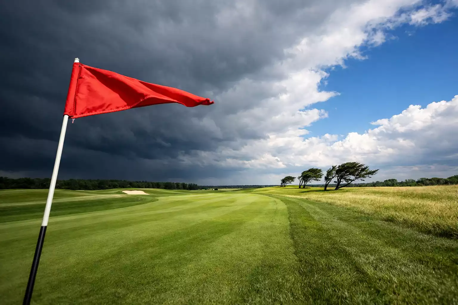 Golf Strategie Wetterfaktor — Windfahne auf einem Golfplatz mit aufziehenden Wolken und Fairway im Hintergrund